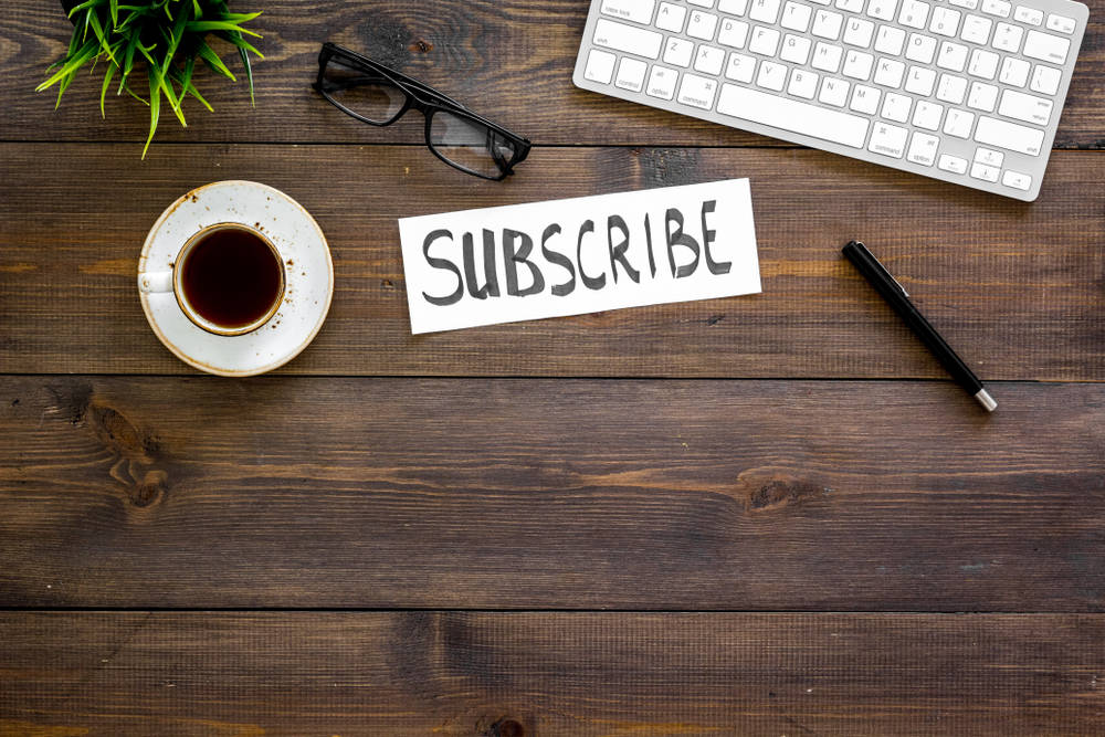 Brown wooden table with a keyboard, coffee mug, pen, glasses, and white paper with the word "subscribe" on it.