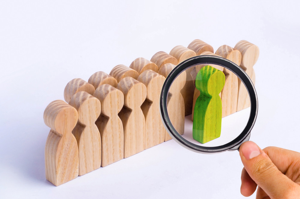 Wooden human figures standing in a line with one painted in green standing in front of the crowd with magnifying glass focused on it.